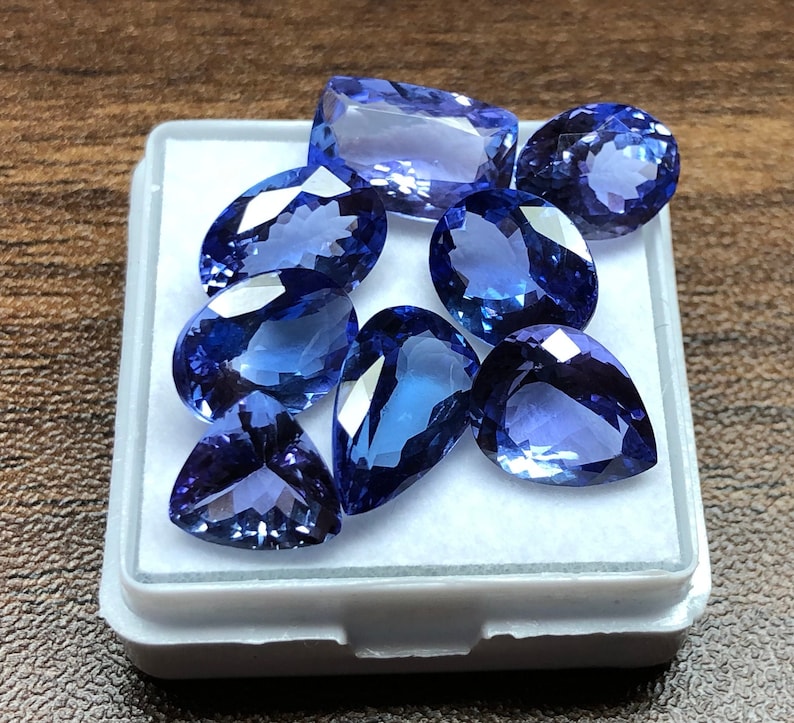 May include: A collection of eight blue pear-shaped gemstones in a white box. The gemstones are various shades of blue and have a smooth, polished finish.