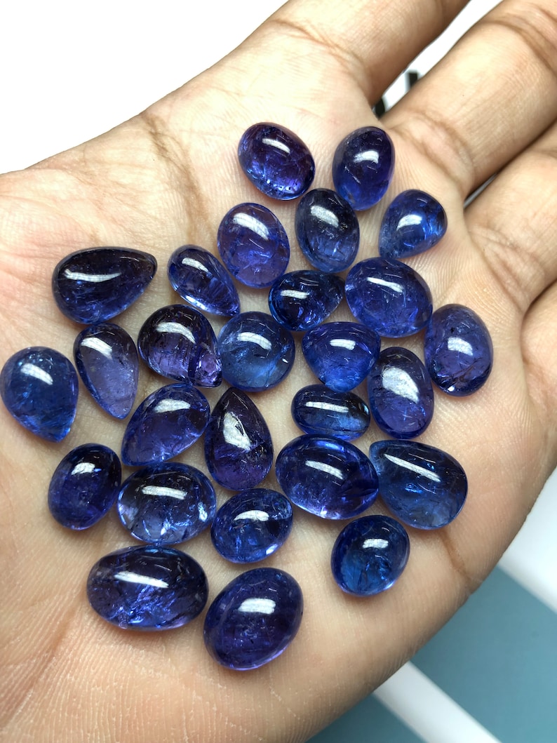 May include: A collection of polished, oval and teardrop-shaped blue gemstones. The gems vary in size and are displayed against a neutral background. The stones have a glossy finish, reflecting light, and are likely used for jewelry making or collecting.