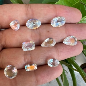 May include: Assortment of iridescent white gemstones in various shapes and sizes, including oval, pear, and square. The gemstones are displayed on a hand.