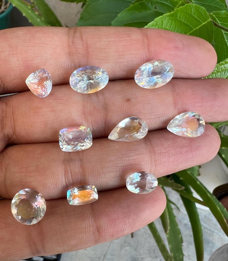 May include: Assortment of iridescent white gemstones in various shapes and sizes, including oval, pear, and square. The gemstones are displayed on a hand.