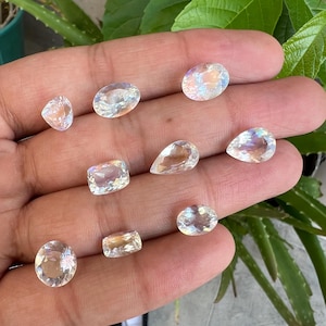 May include: A collection of nine iridescent moonstone gemstones in various shapes and sizes, including oval, pear, and square cuts. The stones are displayed on a hand, showcasing their shimmering, rainbow-like reflections.