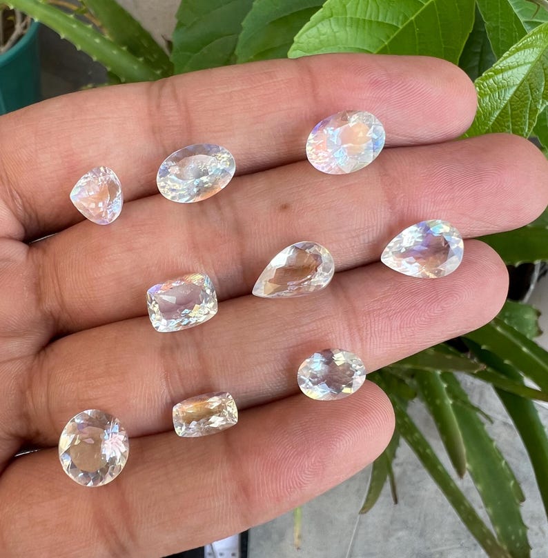 May include: A collection of nine iridescent moonstone gemstones in various shapes and sizes, including oval, pear, and square cuts. The stones are displayed on a hand, showcasing their shimmering, rainbow-like reflections.