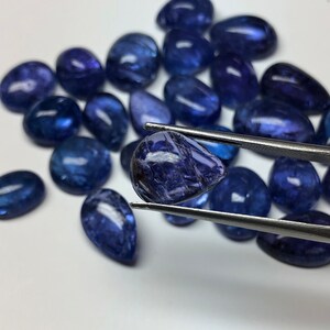 May include: A collection of polished, deep blue gemstones, some teardrop-shaped, others oval. One gem is held by tweezers, showcasing its facets. The gems have a reflective quality, suggesting they are cut and polished for jewelry or display.
