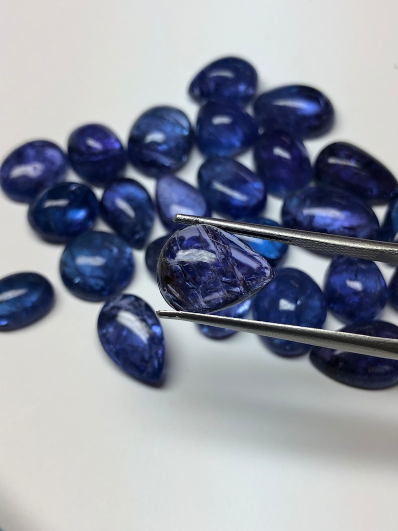 May include: A collection of polished, deep blue gemstones, some teardrop-shaped, others oval. One gem is held by tweezers, showcasing its facets. The gems have a reflective quality, suggesting they are cut and polished for jewelry or display.