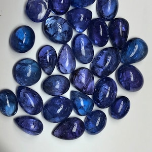 May include: A collection of polished, oval and teardrop-shaped gemstones in varying shades of deep blue and violet. The gemstones are arranged on a white surface, reflecting light and showcasing their smooth, glossy surfaces. The overall theme is jewelry and gemstones.