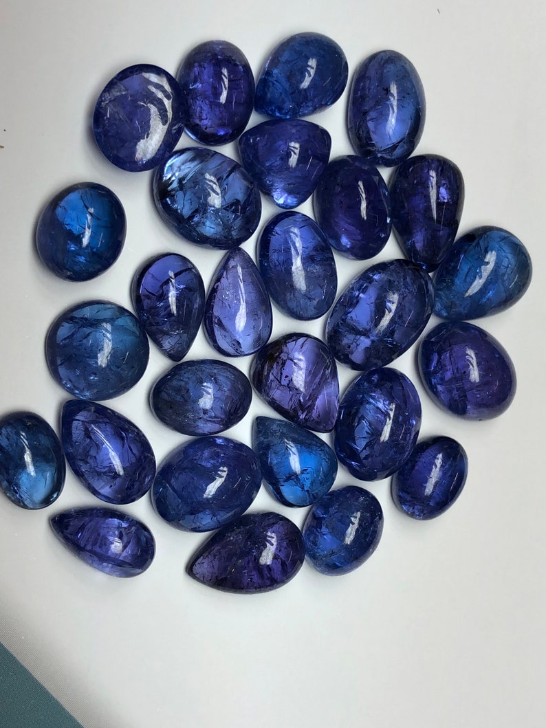 May include: A collection of polished, oval and teardrop-shaped gemstones in varying shades of deep blue and violet. The gemstones are arranged on a white surface, reflecting light and showcasing their smooth, glossy surfaces. The overall theme is jewelry and gemstones.