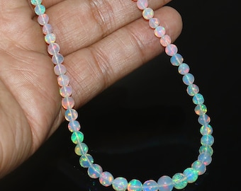 Könnte beinhalten: Eine zarte Halskette aus kleinen, runden Opalperlen. Die Perlen zeigen eine Reihe von Farben, darunter Weiß, Rosa und schillernde Blitze von Grün und Blau. Die Halskette wird vor einem dunklen Hintergrund präsentiert.