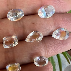 May include: A collection of six iridescent gemstones in various shapes and sizes, including oval, pear, and square. The gemstones are clear and have a rainbow sheen.