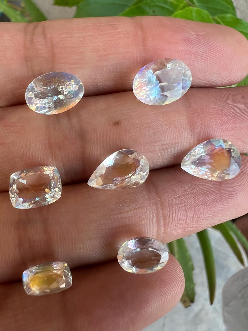 May include: A collection of six iridescent gemstones in various shapes and sizes, including oval, pear, and square. The gemstones are clear and have a rainbow sheen.