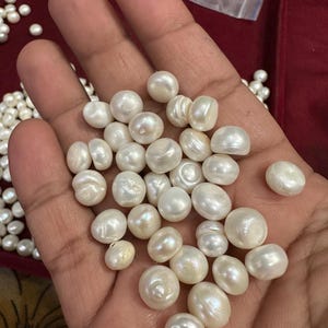 Puede incluir: Primer plano de un puñado de perlas blancas cremosas. Las perlas tienen una forma irregular y un brillo sutil. Se ven más perlas en el fondo, lo que sugiere una selección más amplia de estas gemas orgánicas.