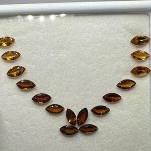 Op de afbeelding: Een verzameling marquise-geslepen edelstenen in tinten amber en bruin, gerangschikt in een gebogen patroon in een witte tray. De edelstenen variëren in kleurintensiteit, van licht goud tot diep roodbruin.