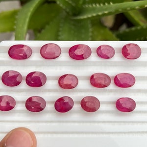 May include: A collection of 15 oval-shaped, faceted ruby gemstones displayed on a white surface. The rubies are a deep red color and have a polished finish.