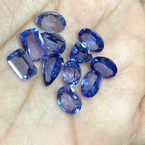 May include: A collection of ten faceted oval and rectangular gemstones in shades of blue and purple. The gemstones are arranged on a hand.