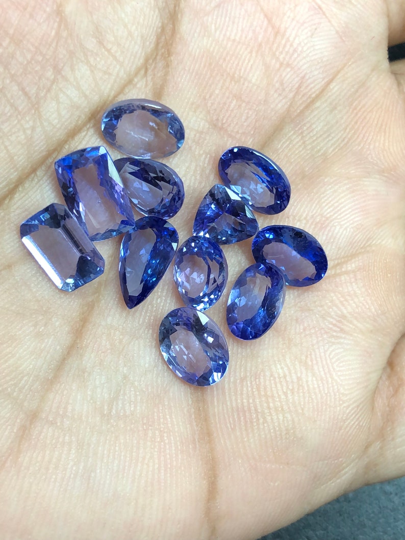 May include: A collection of ten faceted oval and rectangular gemstones in shades of blue and purple. The gemstones are arranged on a hand.