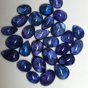 May include: A collection of polished, oval and teardrop-shaped gemstones in shades of blue and purple. The gemstones vary in size and have a translucent quality, with internal patterns visible. The arrangement is scattered on a white surface.