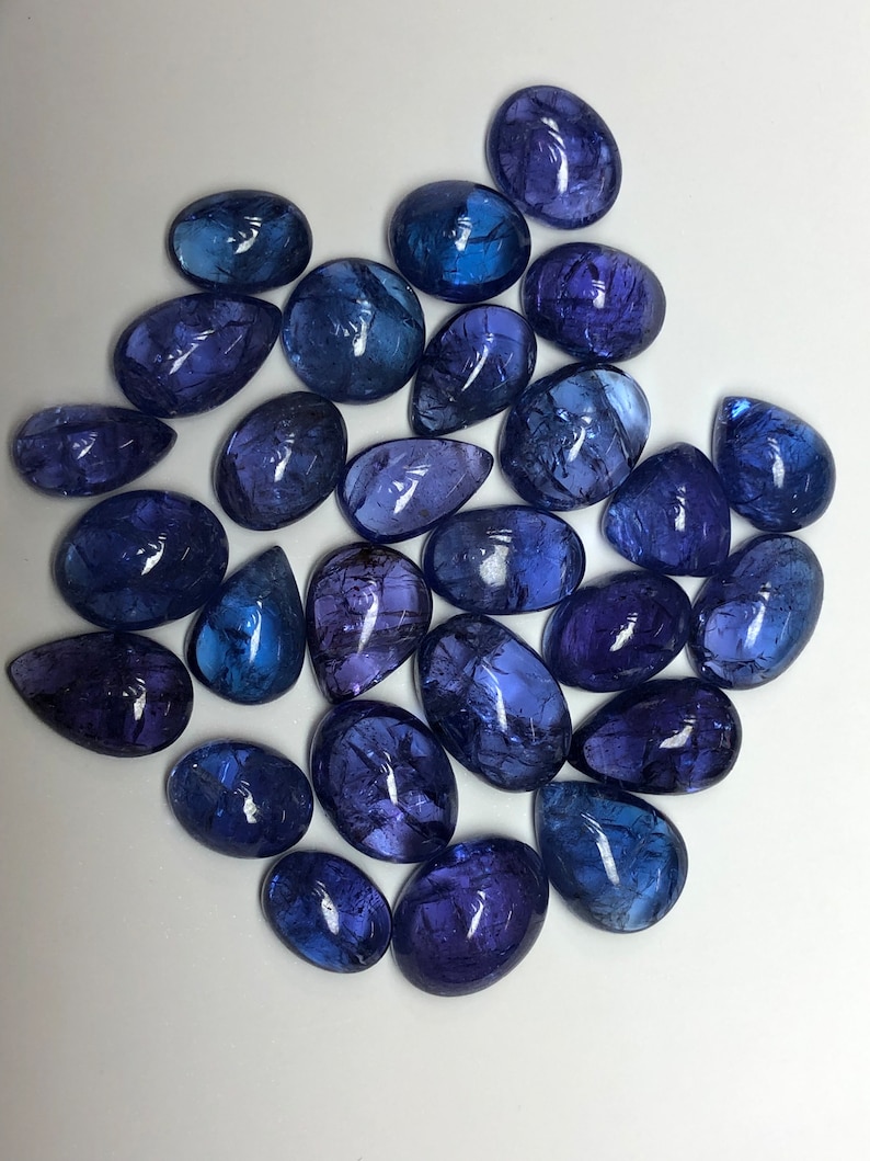 May include: A collection of polished, oval and teardrop-shaped gemstones in shades of blue and purple. The gemstones vary in size and have a translucent quality, with internal patterns visible. The arrangement is scattered on a white surface.