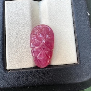 May include: A carved, oval-shaped, ruby-colored gemstone with floral and leaf designs. The gemstone is set in a black and white display case, showcasing the intricate details of the carving. The ruby has a polished, glossy finish.