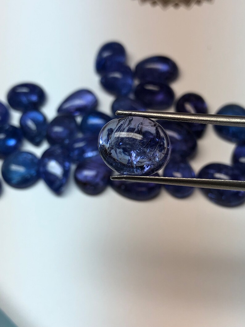 May include: A collection of polished, deep blue gemstones, with one held by tweezers. The gems vary in shape, including oval and teardrop forms. The lighting highlights the internal structures and color variations within the stones.