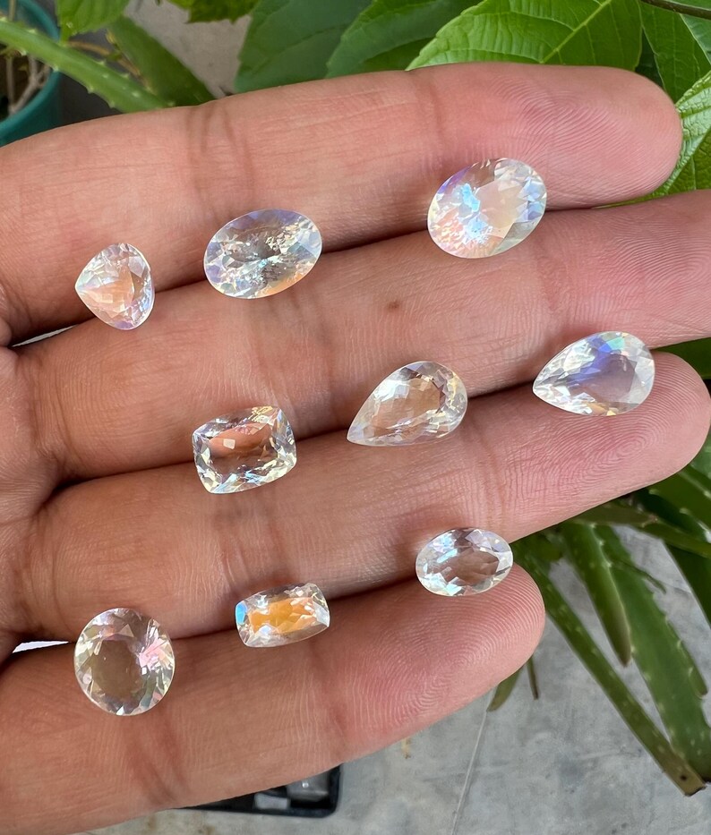 May include: A collection of nine iridescent moonstone gemstones in various shapes and sizes, including oval, pear, and square.