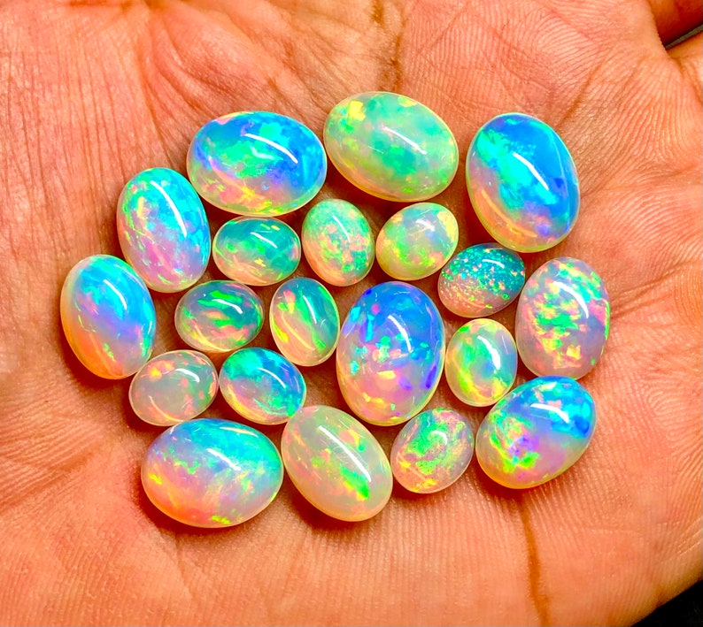 May include: A collection of 17 oval-shaped opal gemstones in various shades of white, blue, green, and pink. The opals are iridescent and have a rainbow-like sheen.