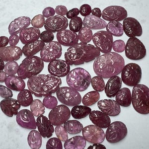 May include: A collection of carved gemstones in varying shapes and sizes, showcasing a range of pink and purple hues. The gemstones feature floral and leaf designs, with intricate details. The arrangement is scattered, highlighting the variety of the collection.