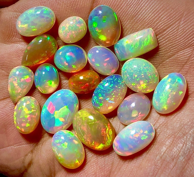 May include: A collection of 17 oval-shaped opals in various shades of white, yellow, and green. The opals are iridescent and have a rainbow-like sheen.