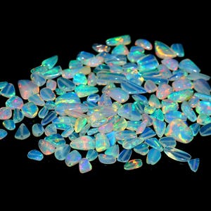 May include: A collection of iridescent opal gemstones, displaying a range of shapes and sizes. The opals exhibit a play of color with flashes of blue, green, and red against a milky white base. The gemstones are scattered on a black background.