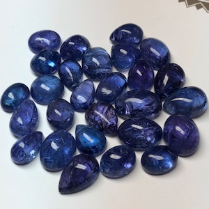 May include: A collection of polished, deep blue tanzanite gemstones. The stones vary in shape, from rounded to teardrop, and display a range of blue hues with subtle internal patterns. The gemstones are arranged on a white surface.