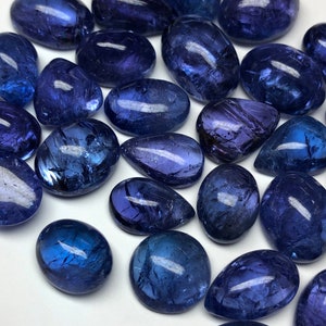 May include: A collection of polished, oval-shaped gemstones in varying shades of deep blue and purple. The stones have a smooth, reflective surface, with natural inclusions visible. The gemstones are scattered across a white surface.