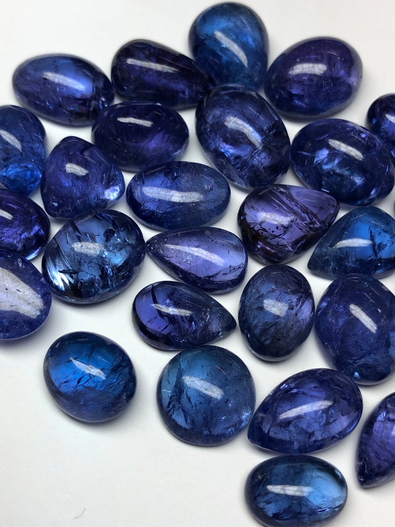 May include: A collection of polished, oval-shaped gemstones in varying shades of deep blue and purple. The stones have a smooth, reflective surface, with natural inclusions visible. The gemstones are scattered across a white surface.