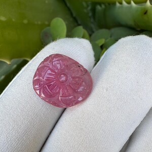 May include: A pink gemstone carved with a floral design. The gemstone is held in a white glove, with green foliage in the background. The carved flower has detailed petals and a central circular element.