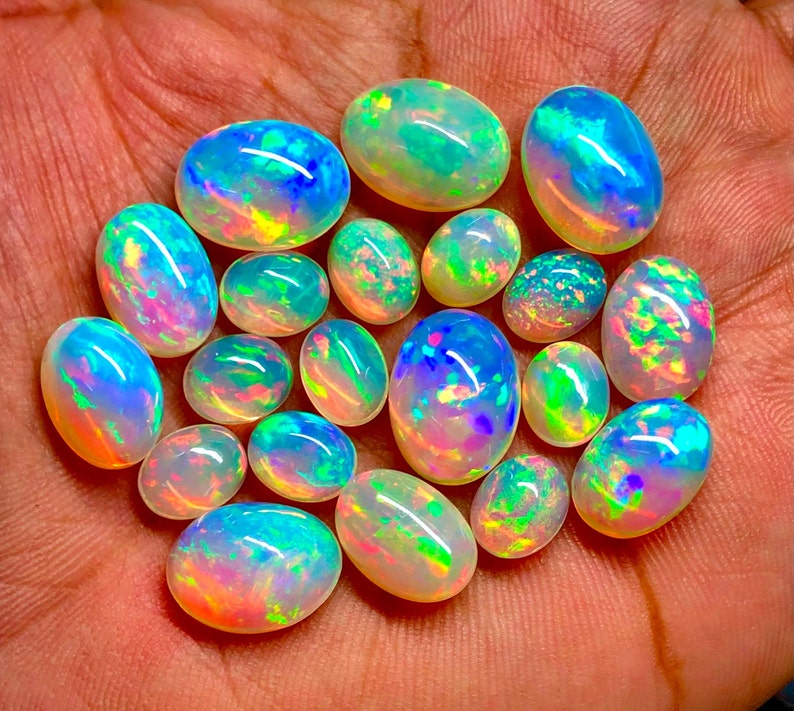 May include: A collection of 17 oval-shaped opals in various shades of white, blue, green, and pink. The opals are iridescent and have a rainbow-like sheen.