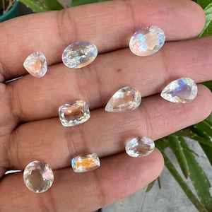 May include: A collection of ten iridescent moonstone gemstones in various shapes and sizes, including oval, pear, and rectangular cuts. The stones are displayed on a hand, showcasing their shimmering, rainbow-like reflections.