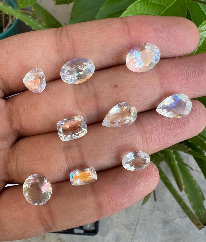 May include: A collection of ten iridescent moonstone gemstones in various shapes and sizes, including oval, pear, and rectangular cuts. The stones are displayed on a hand, showcasing their shimmering, rainbow-like reflections.