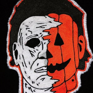 Pode incluir: Patch bordado com um design branco e laranja de um rosto de homem com um rosto de abóbora sobreposto no lado direito do seu rosto.
