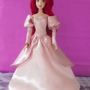 Pode incluir: Um vestido de baile de cetim rosa com mangas bufantes e um decote cora&ccedil;&atilde;o. A boneca est&aacute; usando o vestido e tem cabelo comprido e vermelho e olhos azuis.