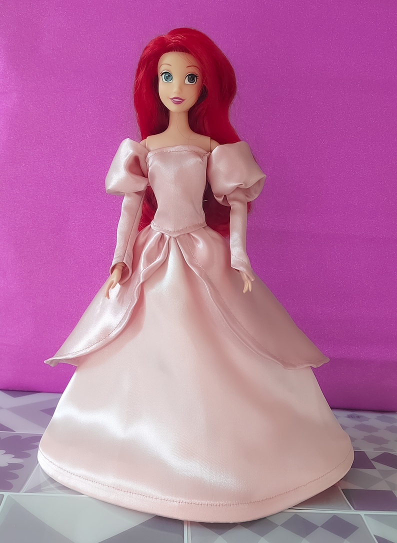 Pode incluir: Um vestido de baile de cetim rosa com mangas bufantes e um decote cora&ccedil;&atilde;o. A boneca est&aacute; usando o vestido e tem cabelo comprido e vermelho e olhos azuis.