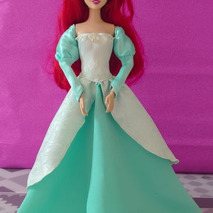 Pode incluir: Uma boneca de cabelo ruivo vestindo um vestido branco e turquesa. A boneca tem um corpete branco com um desenho de concha e uma saia turquesa.