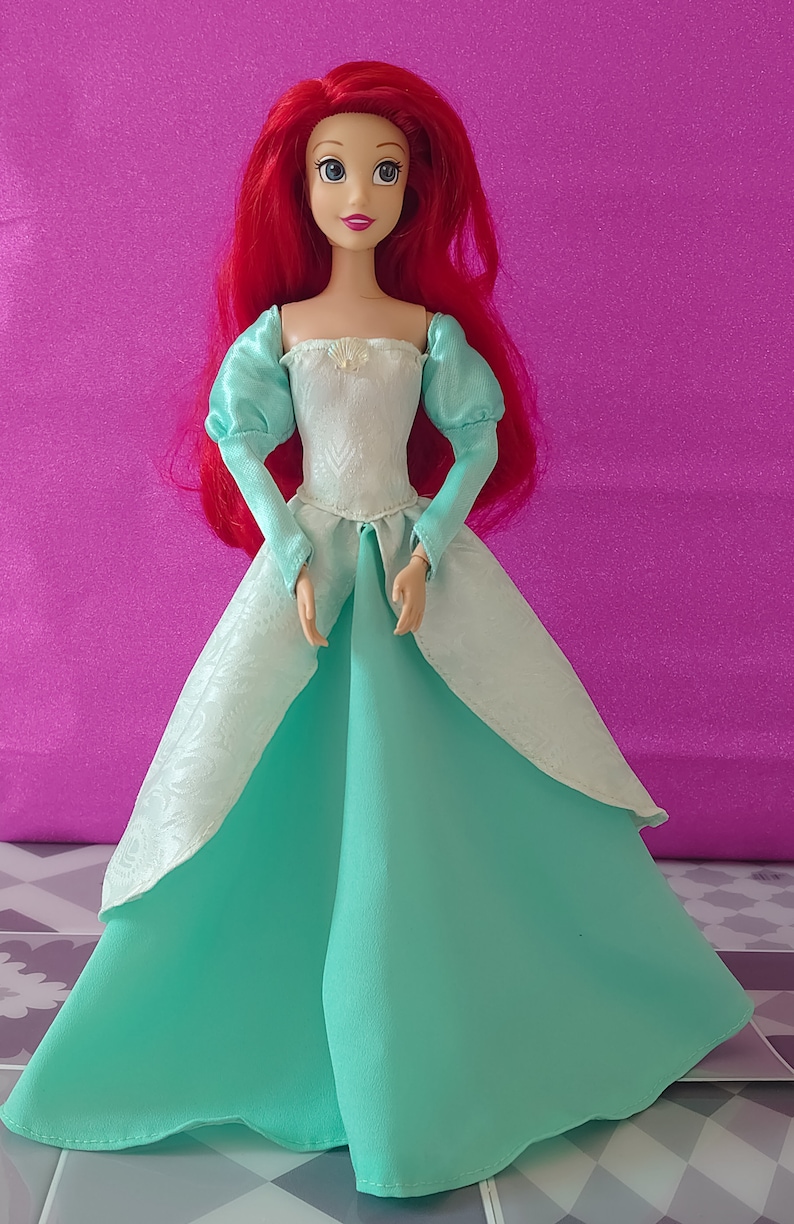 Pode incluir: Uma boneca de cabelo ruivo vestindo um vestido branco e turquesa. A boneca tem um corpete branco com um desenho de concha e uma saia turquesa.