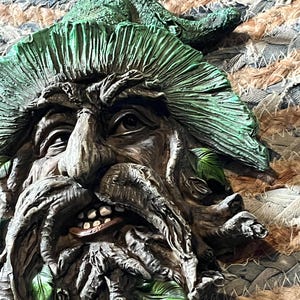 Könnte beinhalten: Detaillierte, dreidimensionale Wandskulptur eines Green Man. Das Gesicht ist aus holzähnlichem Material geschnitzt, mit einem verwitterten, strukturierten Aussehen. Die Skulptur hat einen Bart, Blätterhaare und grüne Akzente.