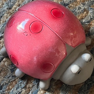 Op de afbeelding: Een keramiek lieveheersbeestje figuur met een rode schil en zwarte stippen. Het lieveheersbeestje heeft een grijs lichaam en zwarte ogen.