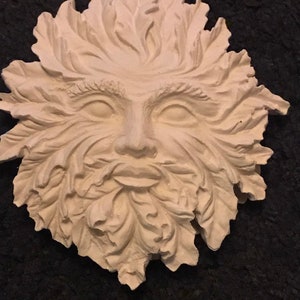 Könnte beinhalten: Eine cremefarbene Green Man-Gesichtsskulptur mit einem detaillierten Design aus Blättern und Gesichtszügen. Die Skulptur hat einen ruhigen Ausdruck und besteht wahrscheinlich aus Gips oder einem ähnlichen Material. Der Hintergrund ist dunkel.