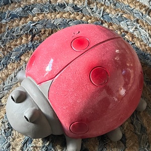 Op de afbeelding: Een rood lieveheersbeestje figuur met zwarte stippen en grijze poten. Het lieveheersbeestje is gemaakt van keramiek en heeft een glanzende afwerking.