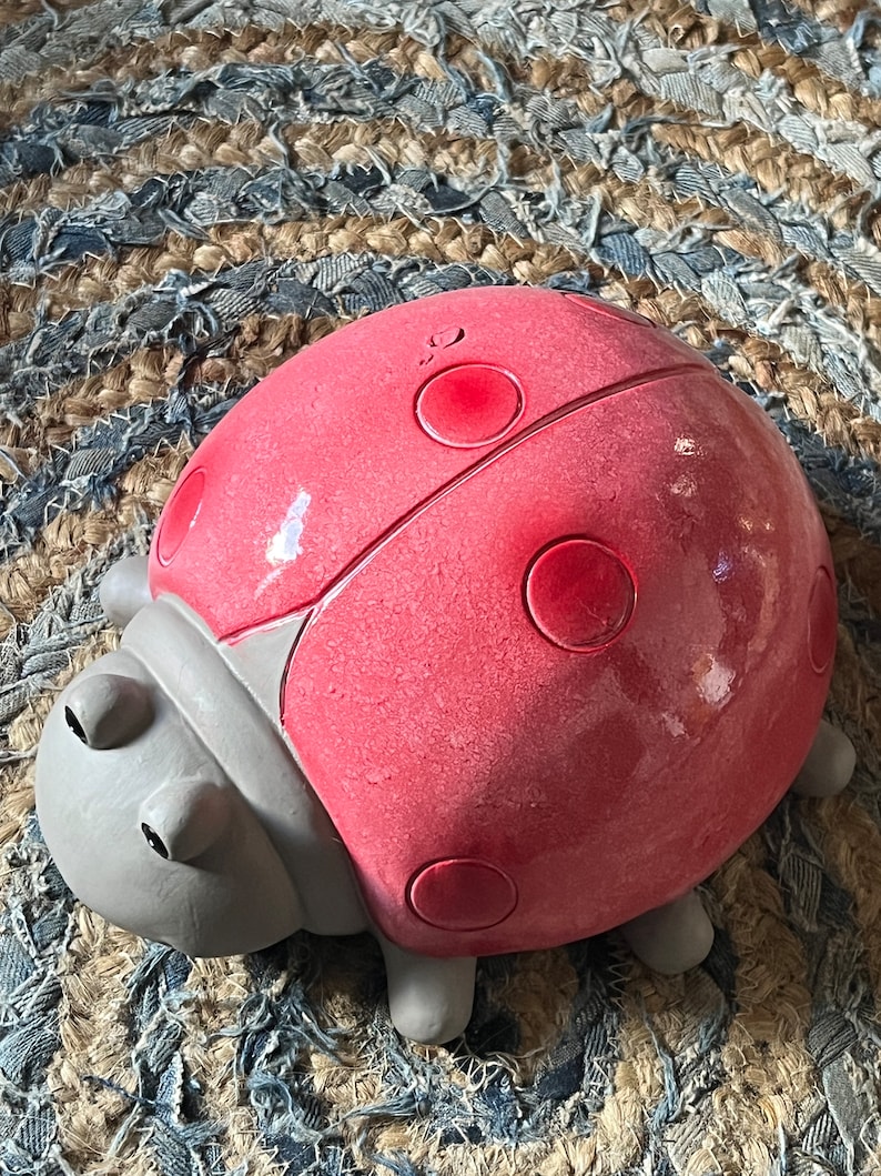Op de afbeelding: Een rood lieveheersbeestje figuur met zwarte stippen en grijze poten. Het lieveheersbeestje is gemaakt van keramiek en heeft een glanzende afwerking.