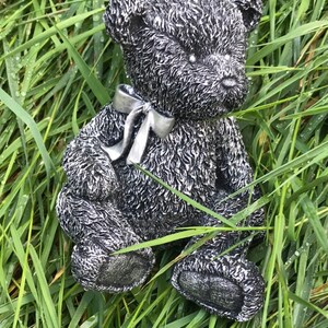 May include: A silver teddy bear figurine with a silver bow sitting in a bed of green grass.
