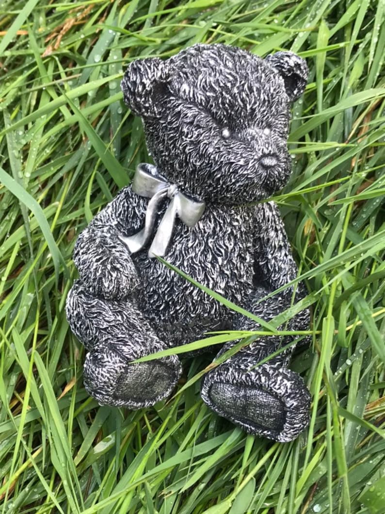 May include: A silver teddy bear figurine with a silver bow sitting in a bed of green grass.