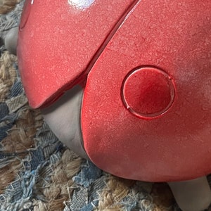 Op de afbeelding: Een rood lieveheersbeestje figuur met een getextureerd oppervlak en een zwarte cirkel op de rug. Het lieveheersbeestje zit op een geweven tapijt.