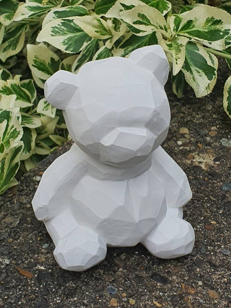 K&ouml;nnte beinhalten: Eine wei&szlig;e geometrische Teddyb&auml;r-Figur. Der B&auml;r sitzt mit gekreuzten Beinen und hat ein schlichtes, modernes Design.