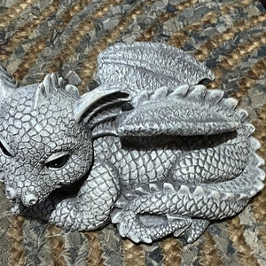May include: A grey dragon figurine with detailed scales, wings, and horns. The dragon is curled up, with its head and wings visible. The figurine is made of a stone-like material, and is a decorative item.