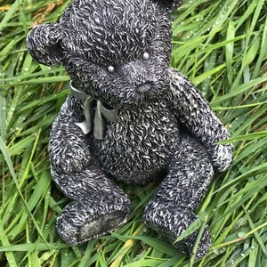 May include: A black and silver teddy bear figurine with a gray ribbon around its neck. The bear is sitting in a bed of green grass.